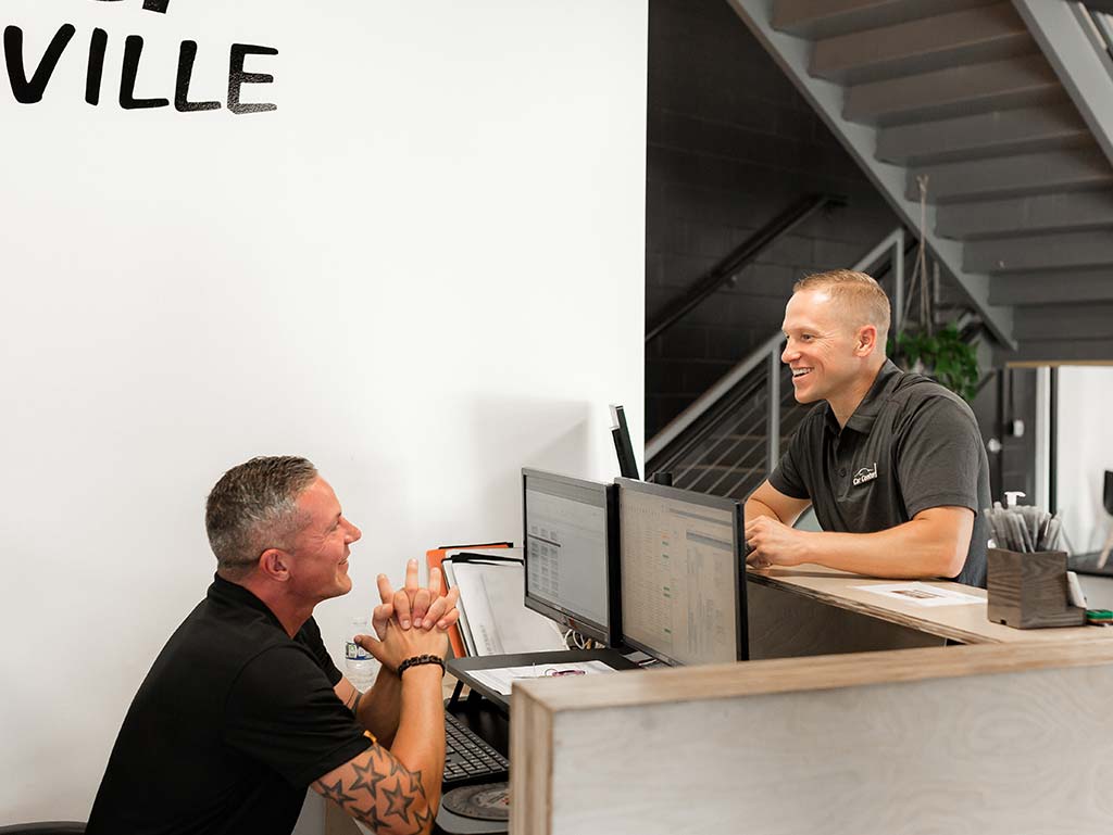 Two men at a reception desk smiling and talking with dual computer monitors and a staircase in the background behind them