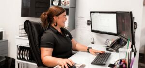 Smiling woman sits at a white desk using a keyboard and mouse with dual computer monitors and office supplies nearby