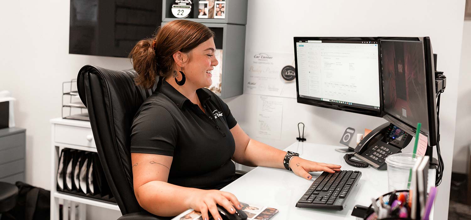 Smiling woman sits at a white desk using a keyboard and mouse with dual computer monitors and office supplies nearby
